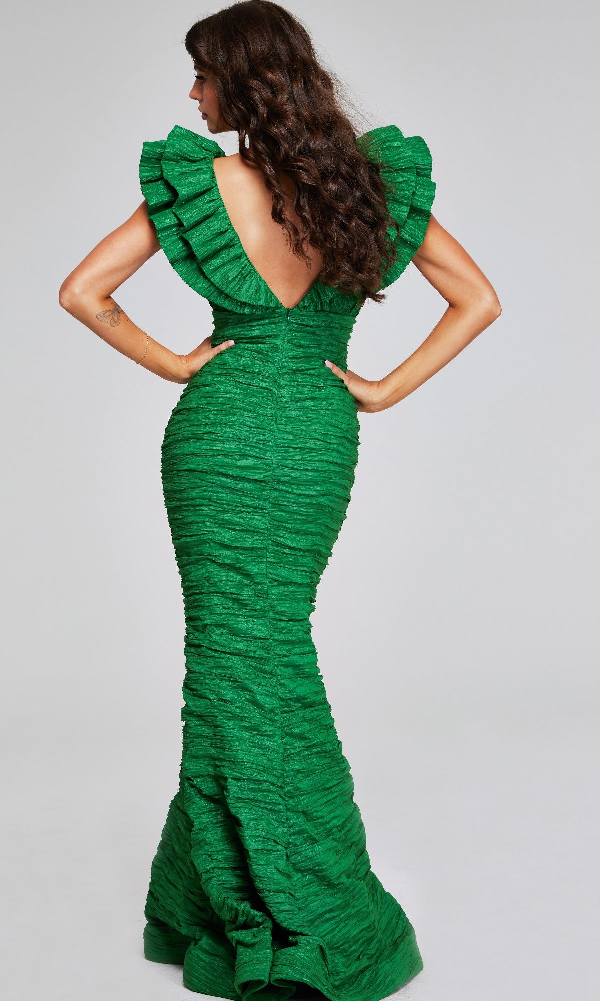 A woman models the Jovani 40044 Long Ruched Formal Dress, featuring a green textured fabric, ruffled sleeves, and a deep V-back as she stands with her back to the camera.